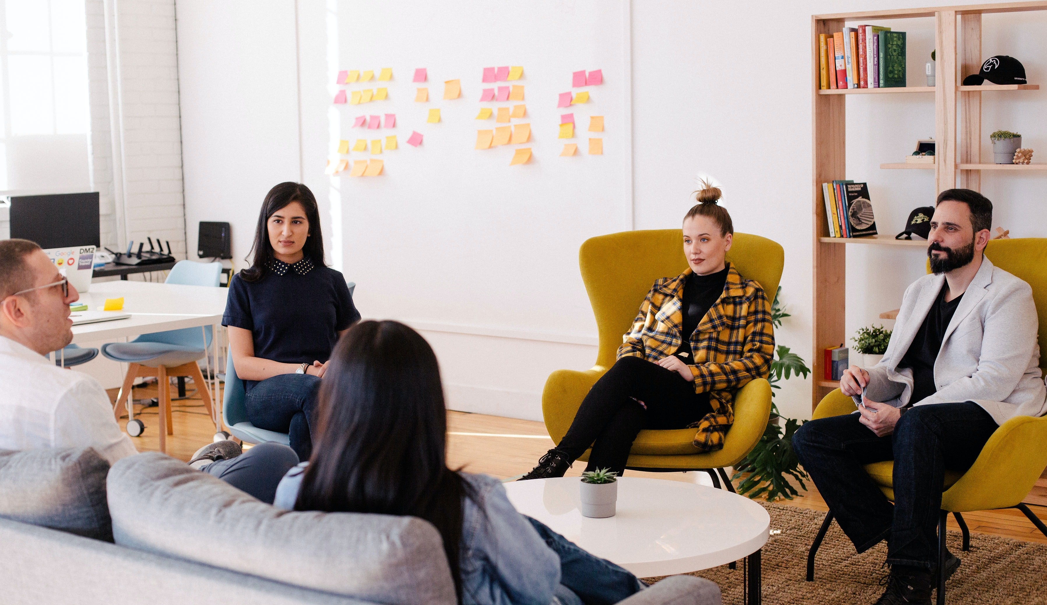 Group of people sitting in chars and sofas, talking. Board with post-its in the background.