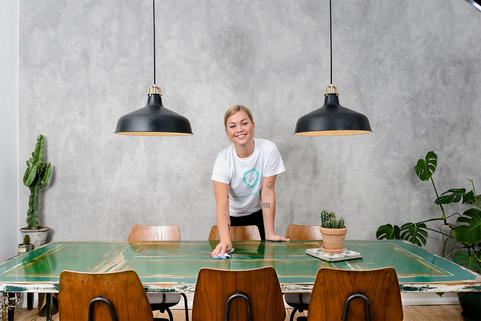 Women looking at the camera while cleaning a table in front of her. Wearing a Happy Helper t-shirt.